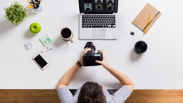 Photographer's overhead workspace with DSLR camera, laptop showing video conference, coffee, and creative accessories.