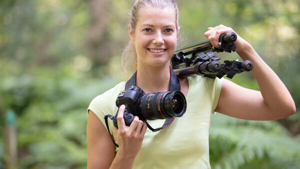 Young woman holding professional video camera and binoculars outdoors among green foliage.