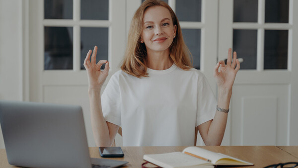 Woman at wooden desk with laptop, notebook, and smartphone, making a playful double peace sign gesture toward camera.