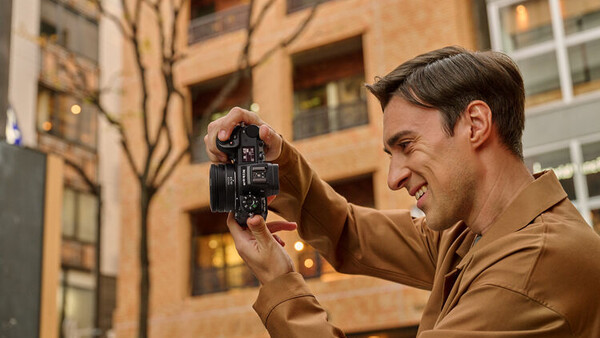 Photographer holding a vintage film camera up to his face, smiling while framing a shot in an urban setting.