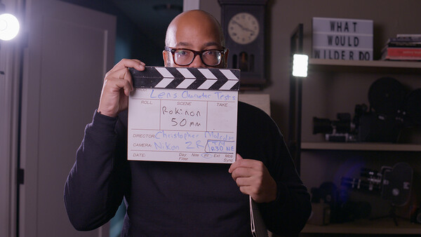 Bald man wearing glasses holds a film clapperboard marked "GARCON SOM" in a home studio setting.