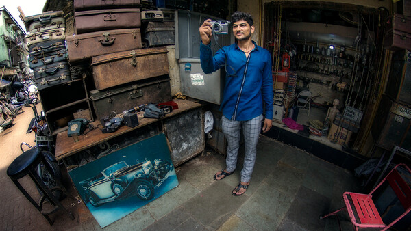 Man in blue jacket holding a camera in a cluttered workshop filled with vintage suitcases and tools.