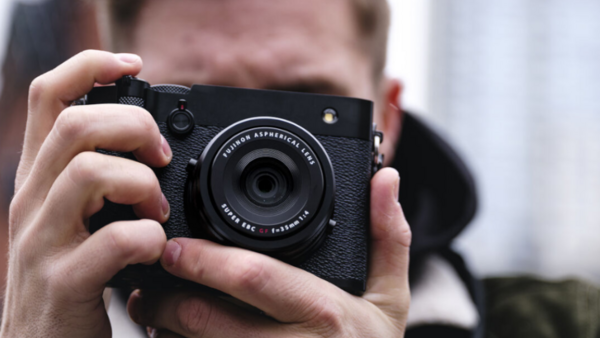 Photographer holding a black rangefinder camera with textured grip, viewed from behind with blurred background.