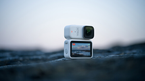 White action camera with flip-out screen displaying ocean footage, positioned on wet rocks with blurred seascape background.