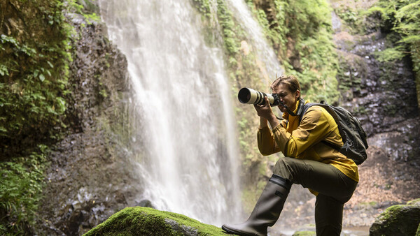 Photographer in yellow jacket crouching by moss-covered rocks, shooting a tall waterfall with a telephoto lens.