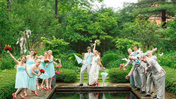 Wedding party posing on either side of a pond with the groom lifting the bride over the water.
