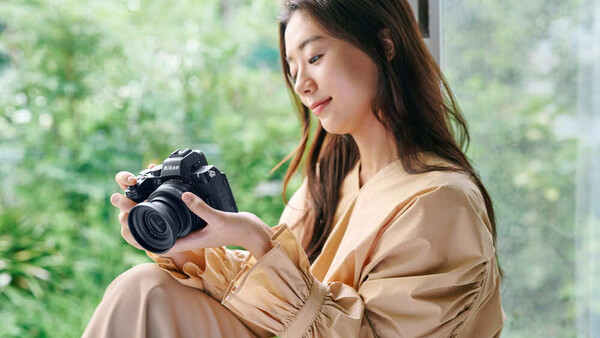 Woman holding a DSLR camera while seated by a bright window with green foliage visible outside.