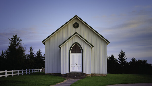 White wooden chapel with peaked roof and arched doorway at dusk, flanked by evergreen trees.