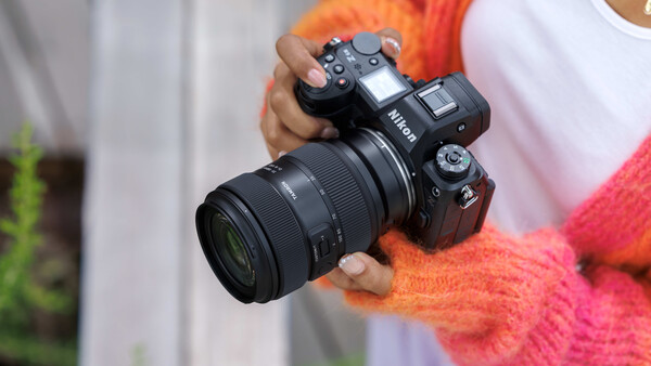Photographer holding a Nikon DSLR camera with telephoto lens while wearing a bright pink and orange knit sweater.