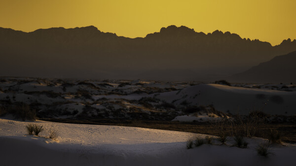 Desert sand dunes silhouetted against a golden sunset sky with mountain ridges in the background.