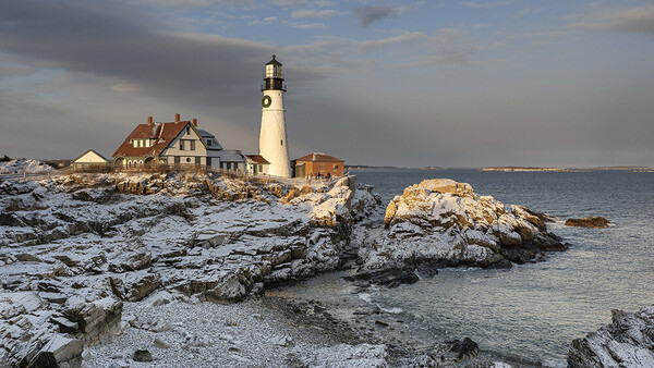 Portland Head Light lighthouse with keeper's house on rocky Maine coastline, snow-covered rocks in foreground, winter conditions.