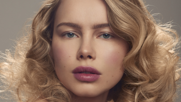 Woman with wavy blonde hair and blue eyes gazing directly at camera against neutral background.