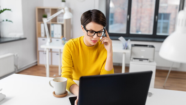 Person in yellow shirt wearing glasses, working at laptop at bright white desk with coffee cup.