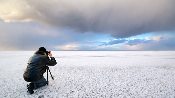 Photographer crouching on a salt flat with camera on tripod, facing dramatic cloudy sky at horizon.