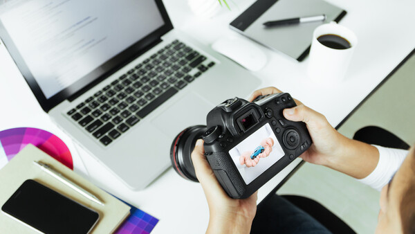 Photographer's hands holding a DSLR camera displaying an image on its rear LCD screen, positioned above a workspace with a laptop and desk accessories.
