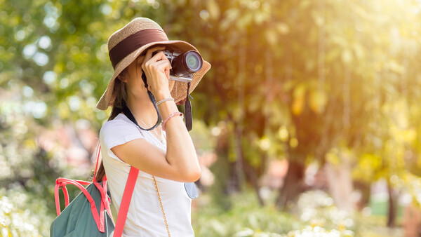 Young person wearing a hat and sunglasses looking through binoculars in a sunlit park setting.
