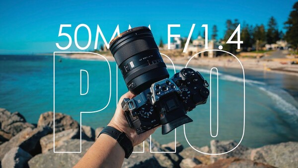 Photographer holding a camera with a 50mm f/1.4 lens at a coastal beach location with clear blue sky.