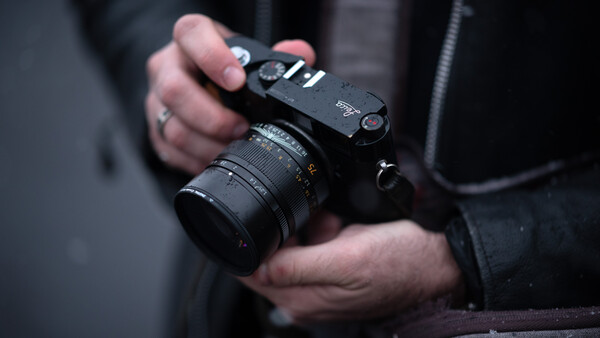 Photographer holding a mirrorless camera with attached lens and shoulder strap.