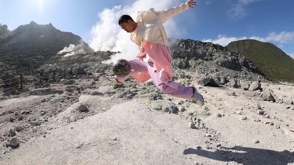 Skateboarder mid-trick on volcanic ash terrain with smoking mountain peaks in background.
