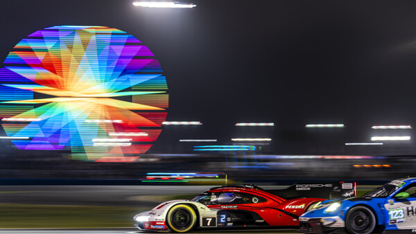 Red and blue race car on track at night with large colorful digital display in background.