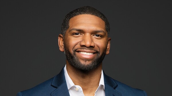 Professional headshot of a man wearing a dark blue blazer and white dress shirt against a dark gray background.