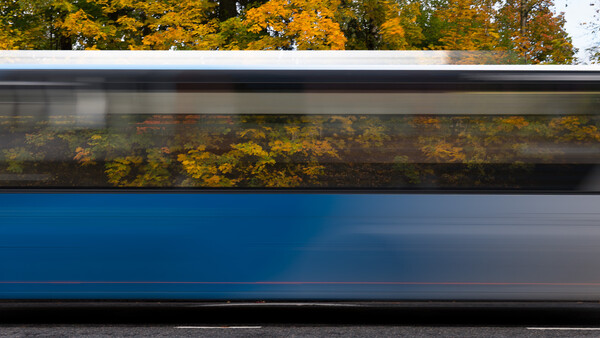Motion-blurred blue bus passing autumn foliage with golden trees reflected in window.
