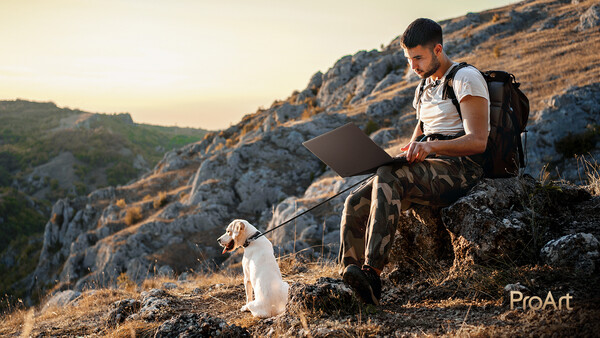 Man sitting on a rocky outcrop working on a laptop with a small dog beside him during golden hour.