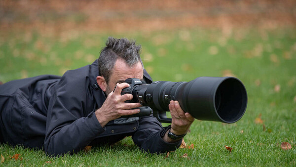 Photographer lying prone on grass while shooting with a telephoto lens.