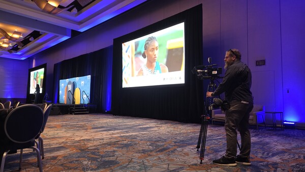 Videographer operating a camera on tripod in a professional event space with blue accent lighting and large projection screens.