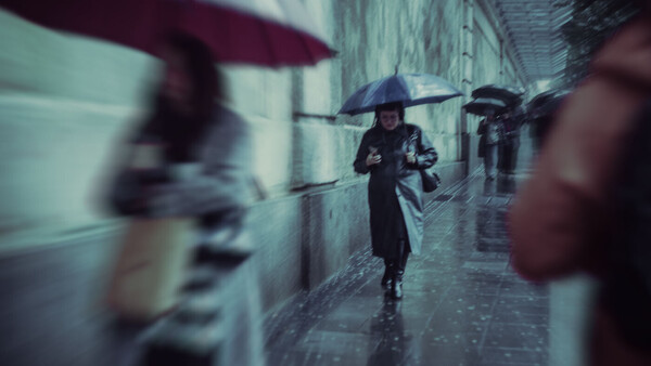 Person with umbrella walking through wet cobblestone alley with blurred figures and buildings