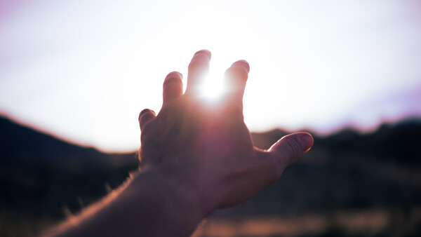 Silhouetted hand reaching toward bright sun against blurred landscape at golden hour
