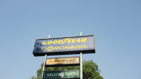 Stacked vintage tire shop signs against a clear blue sky with green tree foliage visible at the base.