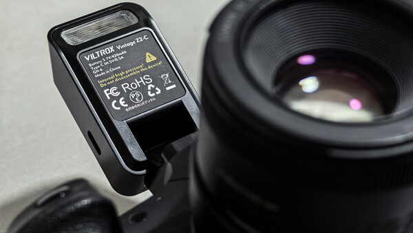 Compact camera flash unit with yellow label mounted on camera body next to telephoto lens.