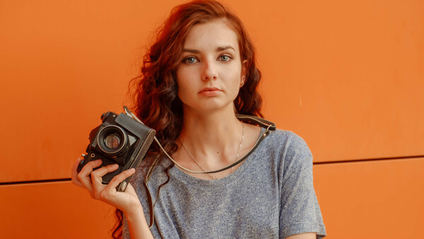 Woman with red wavy hair holding a vintage film camera against an orange background.