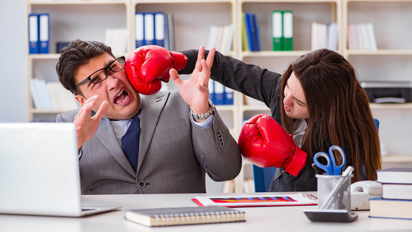 Two business professionals in office attire engage in a playful boxing match at a desk.