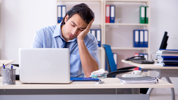 Man in blue shirt leaning on desk, looking tired while working at laptop in office.