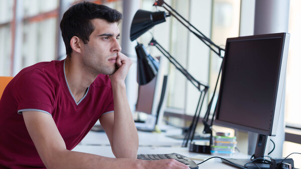 Man in maroon shirt sitting at desk with monitor, appearing thoughtful while working.