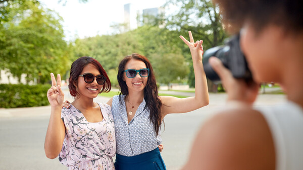 Two women making peace signs while posing for a photo held by a person in the foreground.