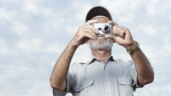 Older man with gray beard and cap holding vintage rangefinder camera up to his face against cloudy sky.