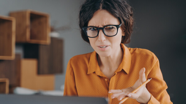 Woman wearing glasses and orange shirt gestures while video conferencing at laptop..