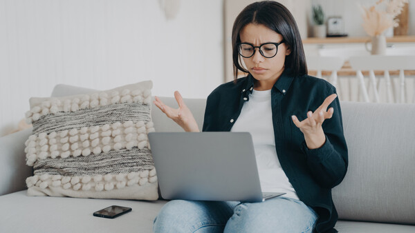 Woman with glasses looking frustrated at laptop screen while sitting on couch.
