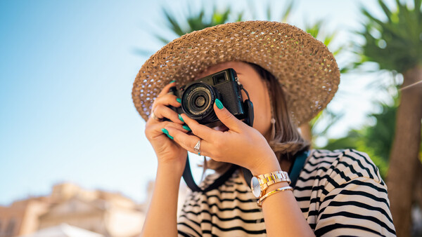Woman holding vintage camera to her face wearing striped shirt and straw hat against tropical backdrop.