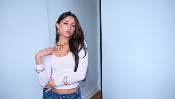 Woman in white cropped long-sleeve top and blue jeans posing against a white wall with geometric shadow.