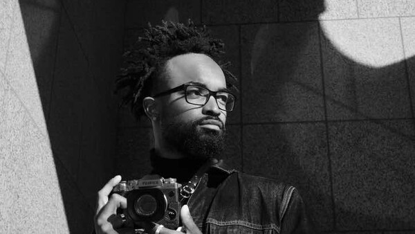 Man wearing glasses and dark jacket holding a camera, photographed against geometric wall shadows in black and white.