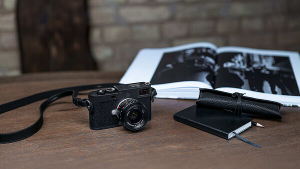 Black rangefinder camera with strap on wooden desk beside photographs and notebook
