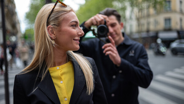 Woman in yellow shirt and black blazer smiling while photographer captures her portrait on an urban street.