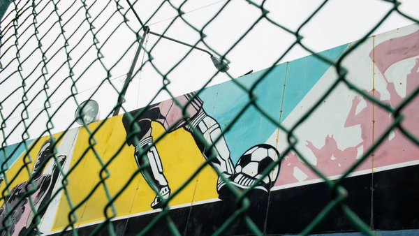 Street art mural of a goalkeeper making a diving save, photographed through chain-link fence