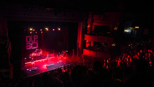 Concert stage bathed in red lighting with band performing to a crowded audience in a dark venue.