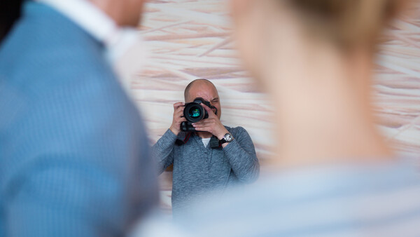 Photographer holding a camera and shooting while two people pose in the foreground, with a brick wall background.