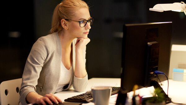 Woman with glasses working at a desk in front of a computer monitor at night.